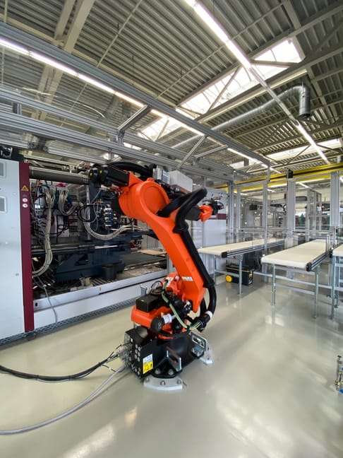 A red and black industrial robotic arm is positioned in a modern factory setting, surrounded by machinery and conveyor belts under bright overhead lights.