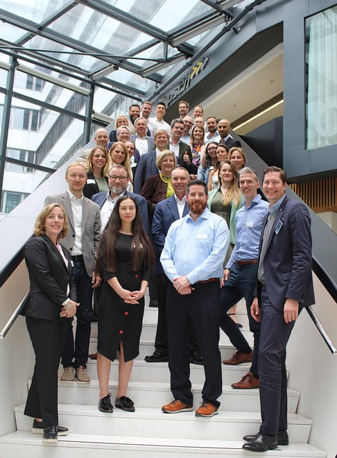 A diverse group of professionals gathers on a staircase in a modern building, smiling for the camera. The group includes men and women of various ages, dressed in business attire, and arranged in multiple rows, showcasing a collaborative atmosphere.