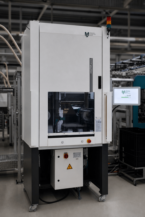 A large, industrial machine with a white exterior, featuring a transparent panel and control screen. It is placed on wheels and is part of a modern factory setting, surrounded by other industrial equipment.