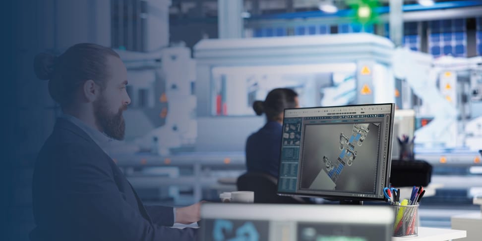 Ein Mann mit Bart sitzt an einem Arbeitsplatz und betrachtet eine detaillierte digitale Grafik auf seinem Computerbildschirm. Im Hintergrund arbeitet eine weitere Person an einem anderen Computer in einer modernen, industriellen Umgebung.