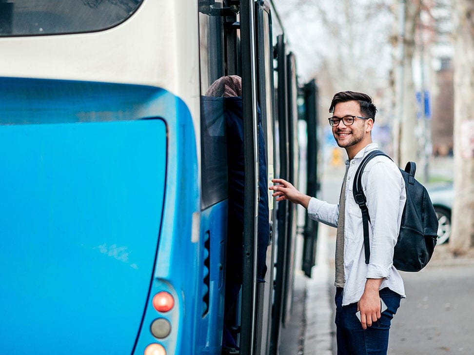 A young man with glasses and a backpack smiles as he prepares to board a blue bus. The background features trees and a city street.